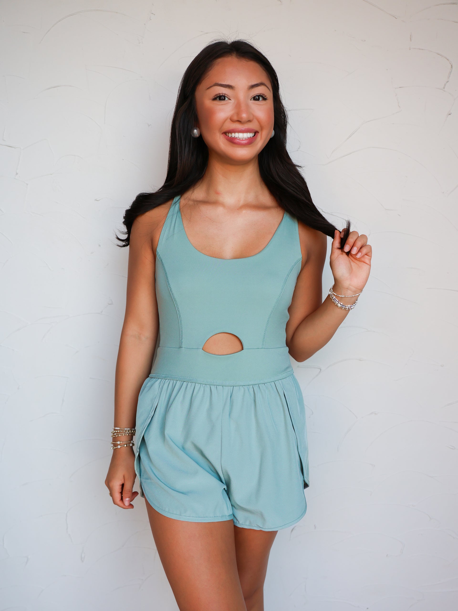 A mid-shot photograph of a smiling brunette woman posing while holding a strand of her hair against a textured white wall. She is wearing a sage green one-piece activewear romper made from a 76% nylon and 24% spandex blend fabric, featuring tank straps, a scoop neckline, a small circular front cutout at the midriff, and attached running shorts with dolphin hemlines