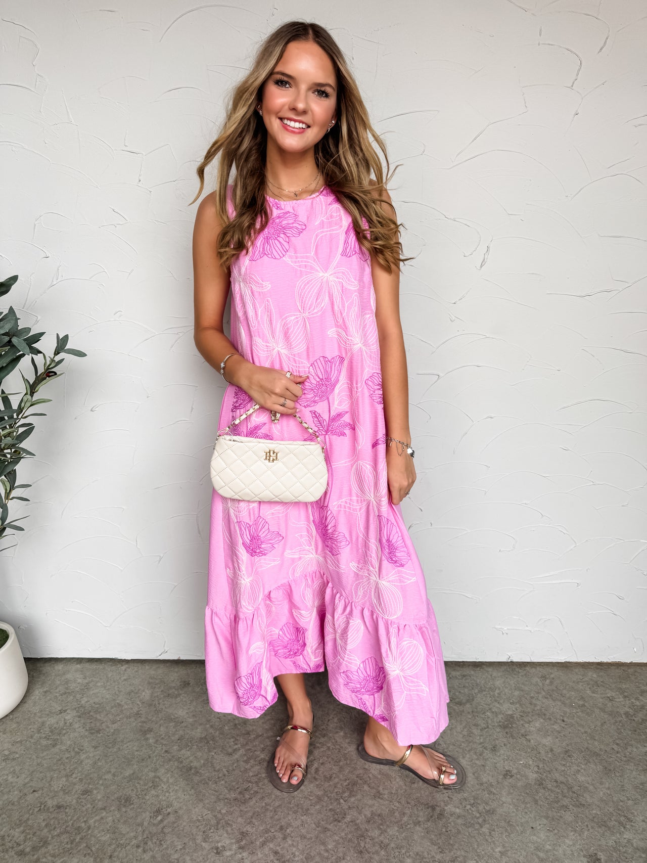 A full-length photograph of a smiling blonde woman posing against a textured white wall while holding a small cream quilted chain handbag. She is wearing a sleeveless pink maxi dress featuring a large white and purple floral print made from 100% polyester fabric, designed with a crew neckline and a ruffled tiered hem, styled with flat sandals.