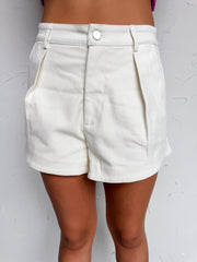A close-up photograph focusing from the waist down of a woman wearing high-waisted, off-white denim shorts made from a 97% cotton and 3% spandex blend against a textured white wall. The shorts feature a structured front pleated design, a silver front button closure, and deep side slash pockets.