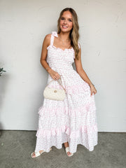A full-length photograph of a smiling blonde woman posing against a textured white wall while holding a small cream quilted chain handbag. She is wearing a white maxi dress featuring a pink ditsy floral print made from 100% polyester fabric, designed with a form-fitting smocked bodice, adjustable tie-shoulder straps, and a tiered ruffle skirt, styled with white flat sandals.
