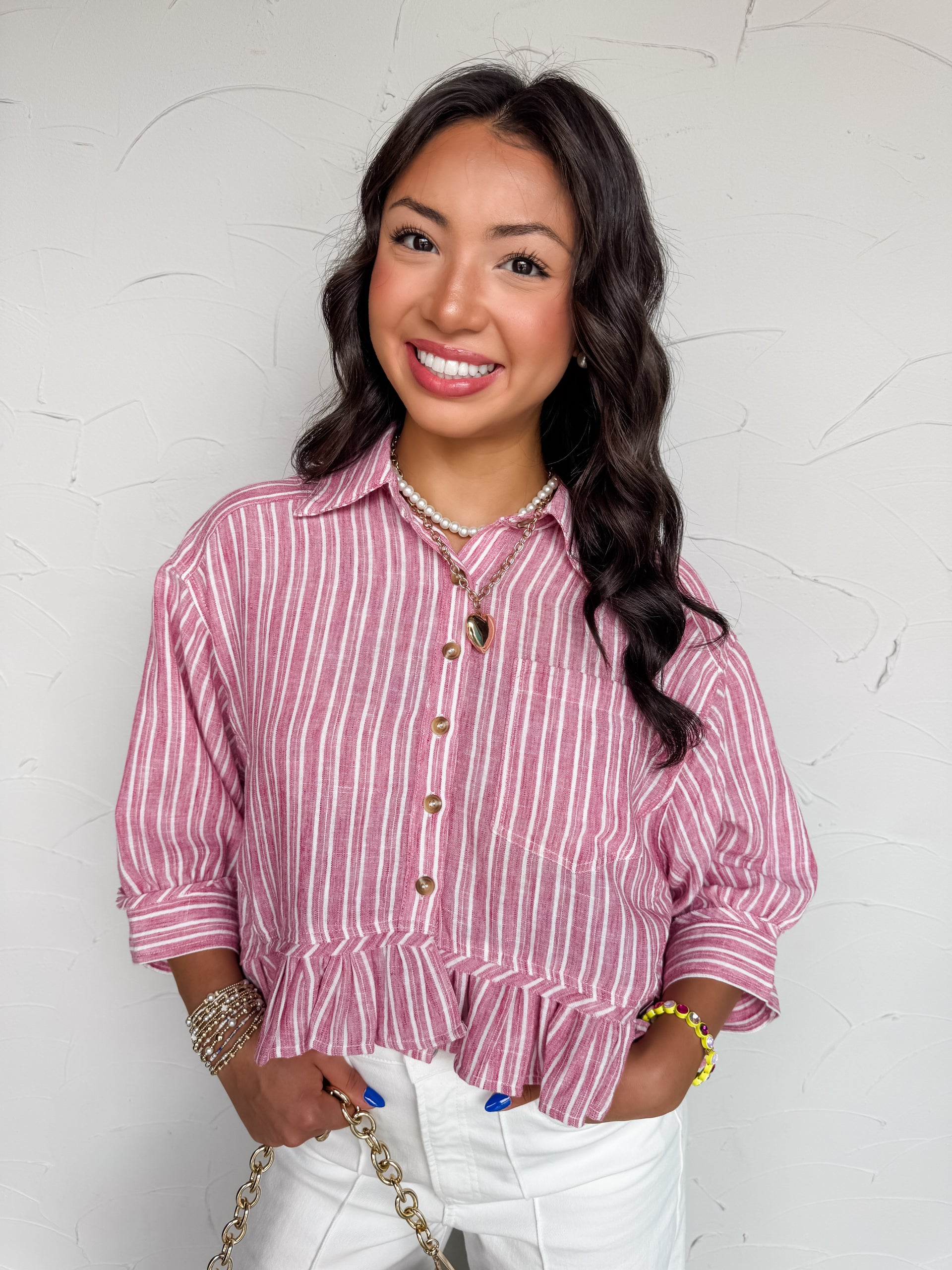 A mid-shot photograph of a smiling brunette woman posing against a textured white wall with her hands in her pockets. She is wearing a mauve and white vertically striped button-down top made from 100% polyester fabric, featuring a pointed collar, a single chest pocket, and a ruffled peplum hemline, styled with white pants.