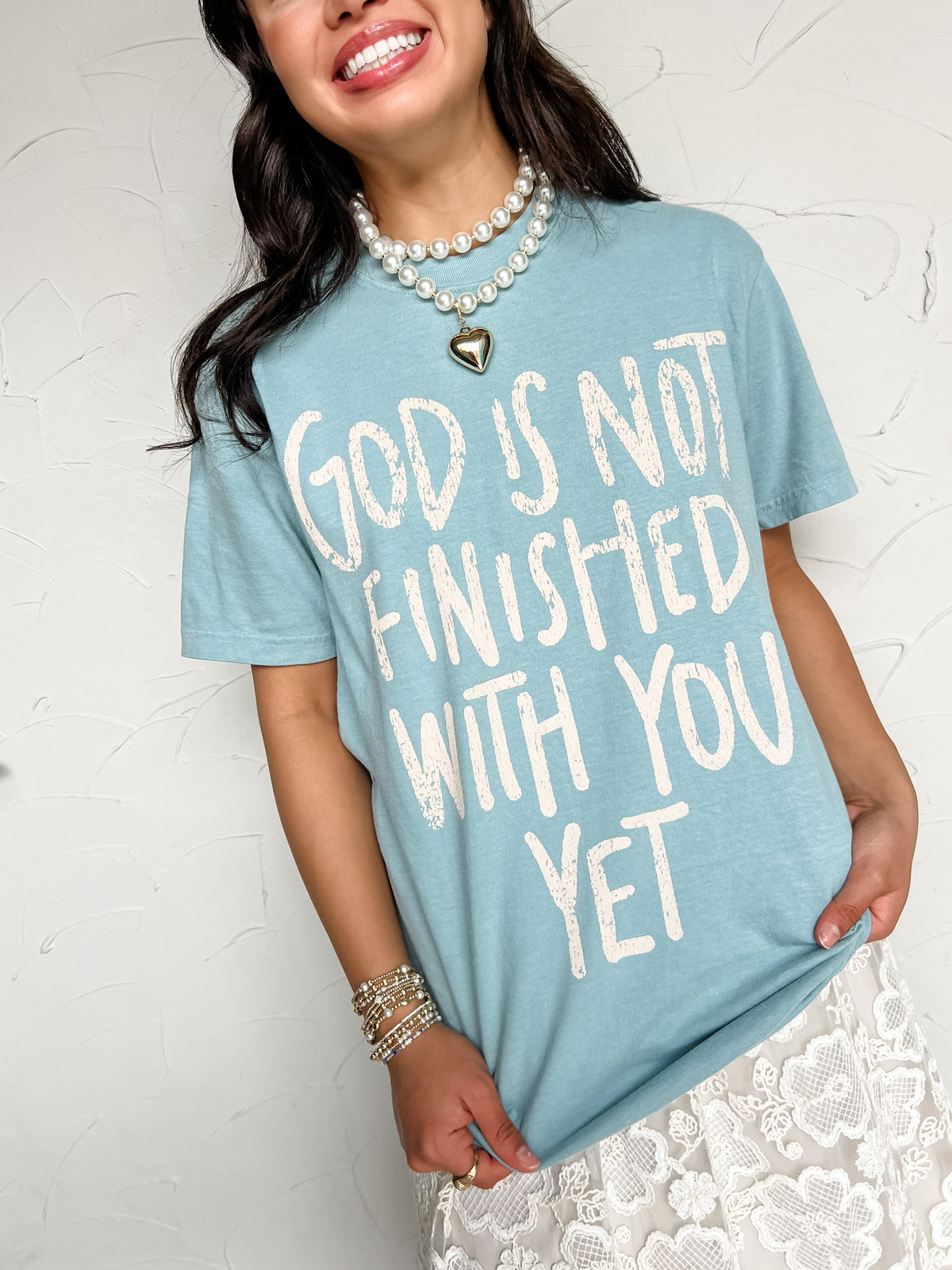 A woman wearing a light blue graphic t-shirt with the phrase 'GOD IS NOT FINISHED WITH YOU YET' in white distressed lettering, styled with a white lace skirt and pearl necklaces.