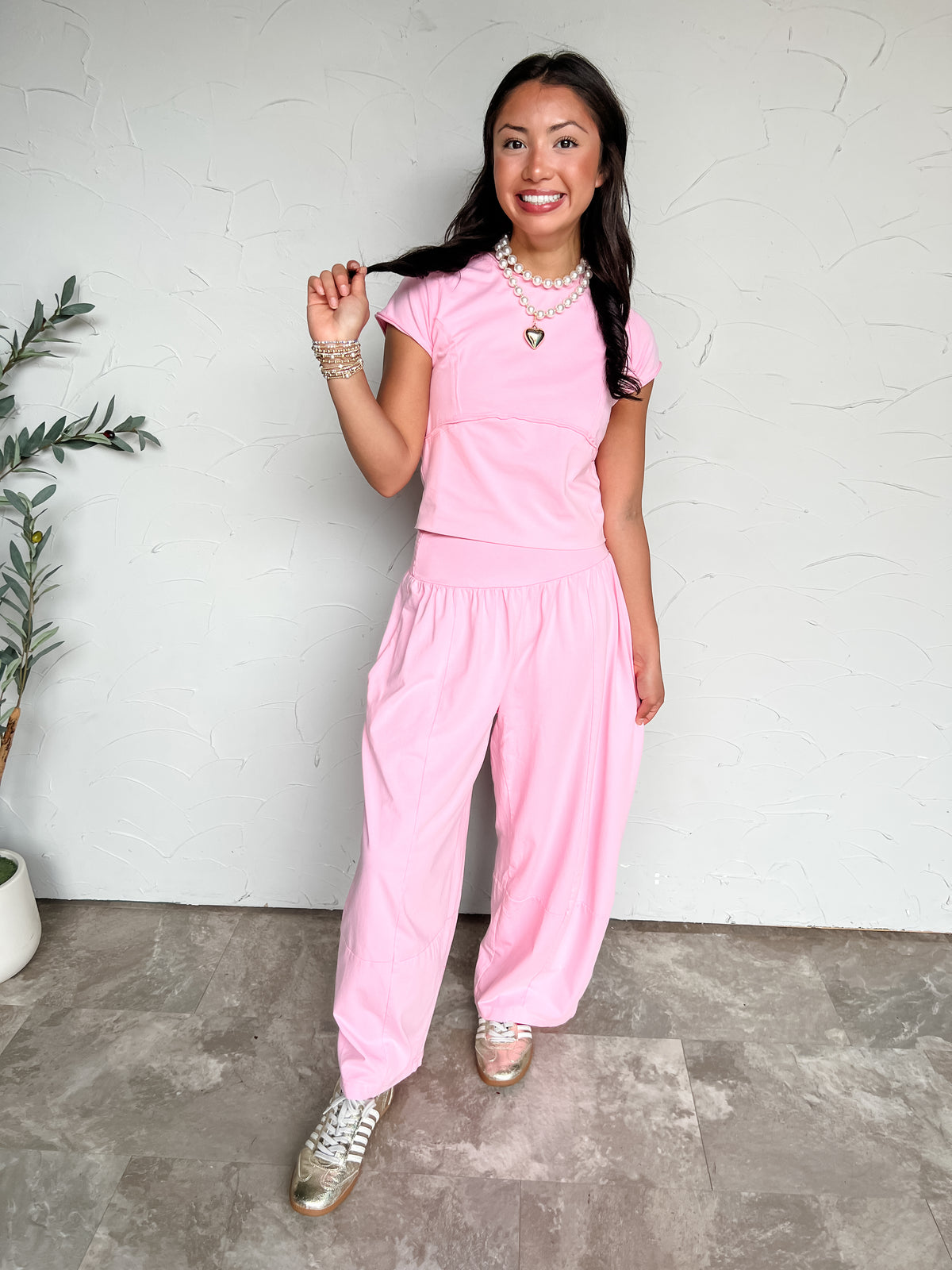 A full-length photograph of a smiling brunette woman with long dark wavy hair holding a strand of her hair while posing against a textured off-white wall next to a potted olive plant. She is wearing a light pink two-piece set made from a 95% cotton and 5% spandex blend, featuring a fitted short cap-sleeved tee with horizontal seam detailing and matching loose wide-leg pants with a wide waistband and panel seams, styled with metallic gold sneakers, a double-strand pearl necklace with a gold puffy heart penda