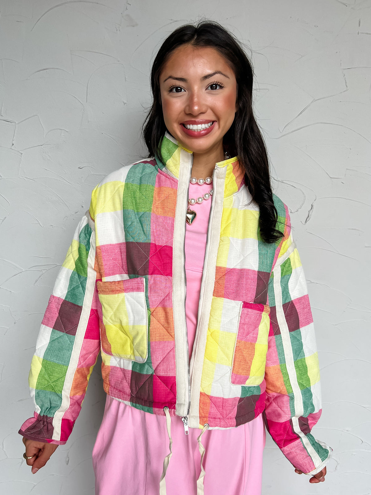 A portrait photograph of a smiling brunette woman with long dark wavy hair posing against a textured off-white wall. She is wearing a colorful quilted zip-up jacket made from a 97% polyester and 3% spandex blend, featuring a vibrant multicolor plaid pattern in pink, yellow, green, and orange, a high stand collar, cream trim along the full zip front, front flap patch pockets, and an adjustable drawstring hem, layered over a bright pink dress and styled with a white pearl necklace featuring a gold heart penda
