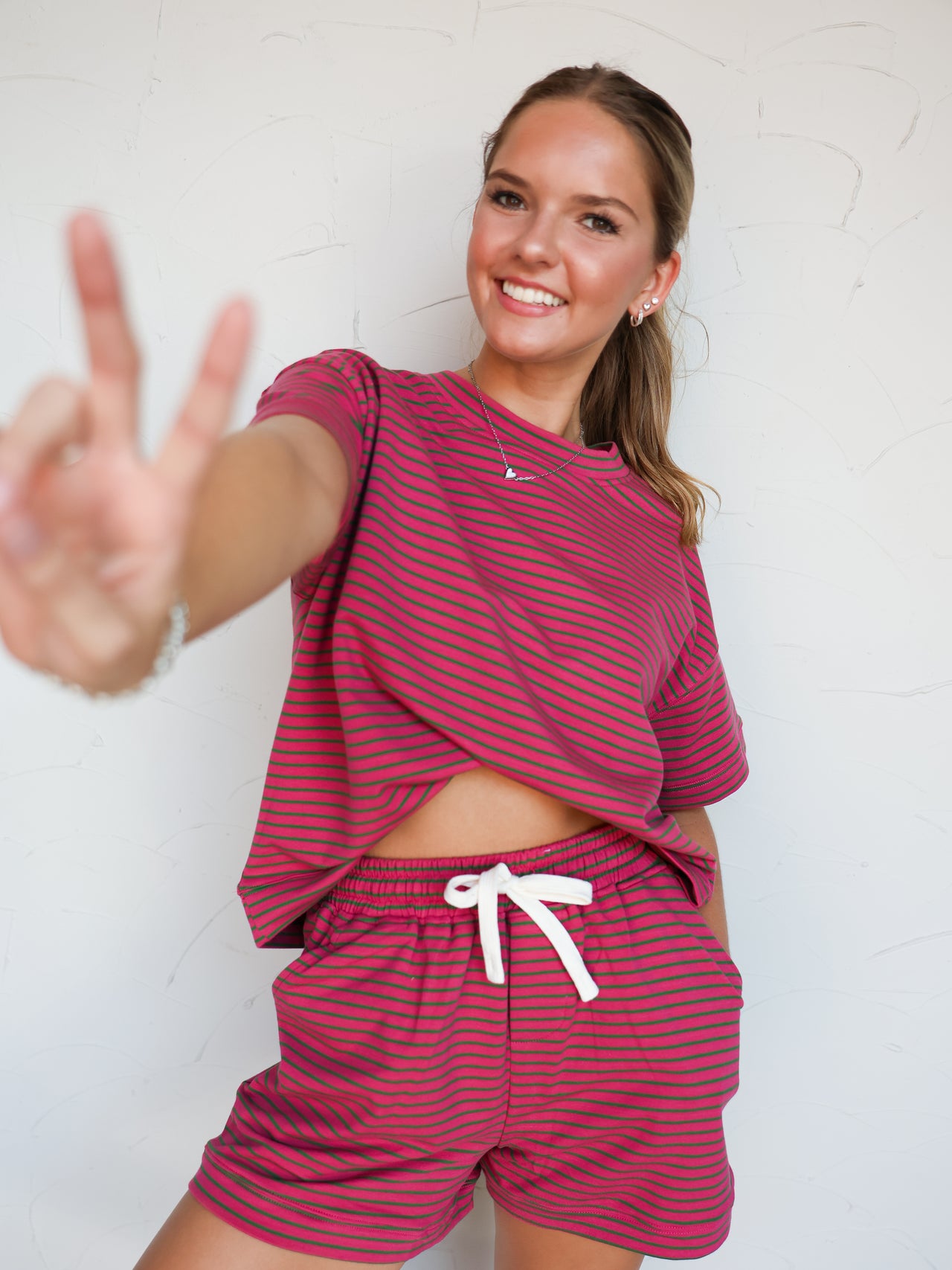 A mid-shot photograph of a smiling blonde woman holding up a peace sign toward the camera against a textured white wall. She is wearing a matching lounge set made from a 93% cotton and 7% spandex blend fabric featuring dark pink and green horizontal stripes, consisting of a relaxed drop-shoulder short-sleeve t-shirt and coordinating drawstring shorts.