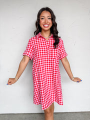 A mid-shot photograph of a smiling brunette woman posing against a textured white wall. She is wearing a short-sleeve shirt dress made from a 50% cotton and 50% polyester blend fabric featuring a red and pink gingham check pattern, a pointed collar with a front button placket, and a relaxed shift silhouette.