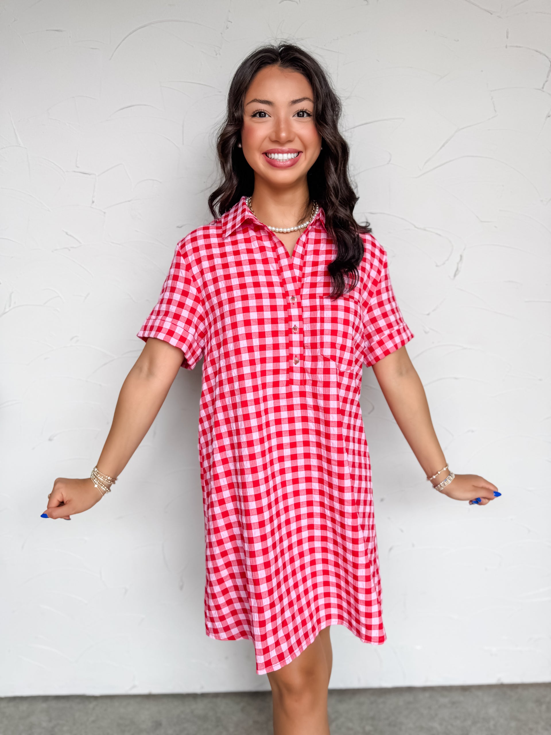 A mid-shot photograph of a smiling brunette woman posing against a textured white wall. She is wearing a short-sleeve shirt dress made from a 50% cotton and 50% polyester blend fabric featuring a red and pink gingham check pattern, a pointed collar with a front button placket, and a relaxed shift silhouette.