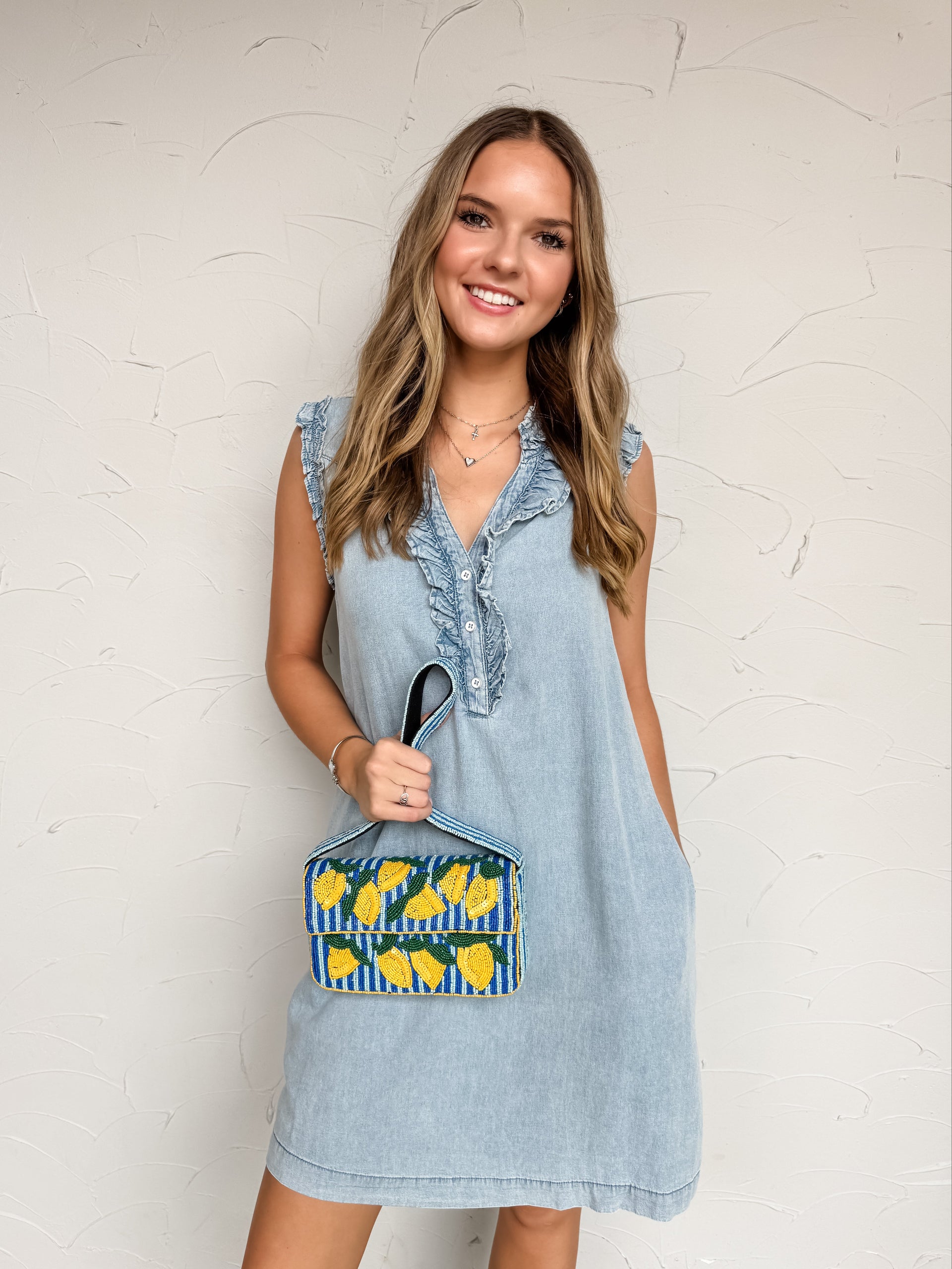 A mid-shot photograph of a smiling blonde woman posing against a textured white wall while holding a blue and yellow beaded lemon-patterned handbag. She is wearing a light-wash denim shift dress made from a soft 65% cotton and 35% Tencel blend fabric, featuring a partial front button placket, cascading ruffle details along the neckline, and small ruffled sleeves.