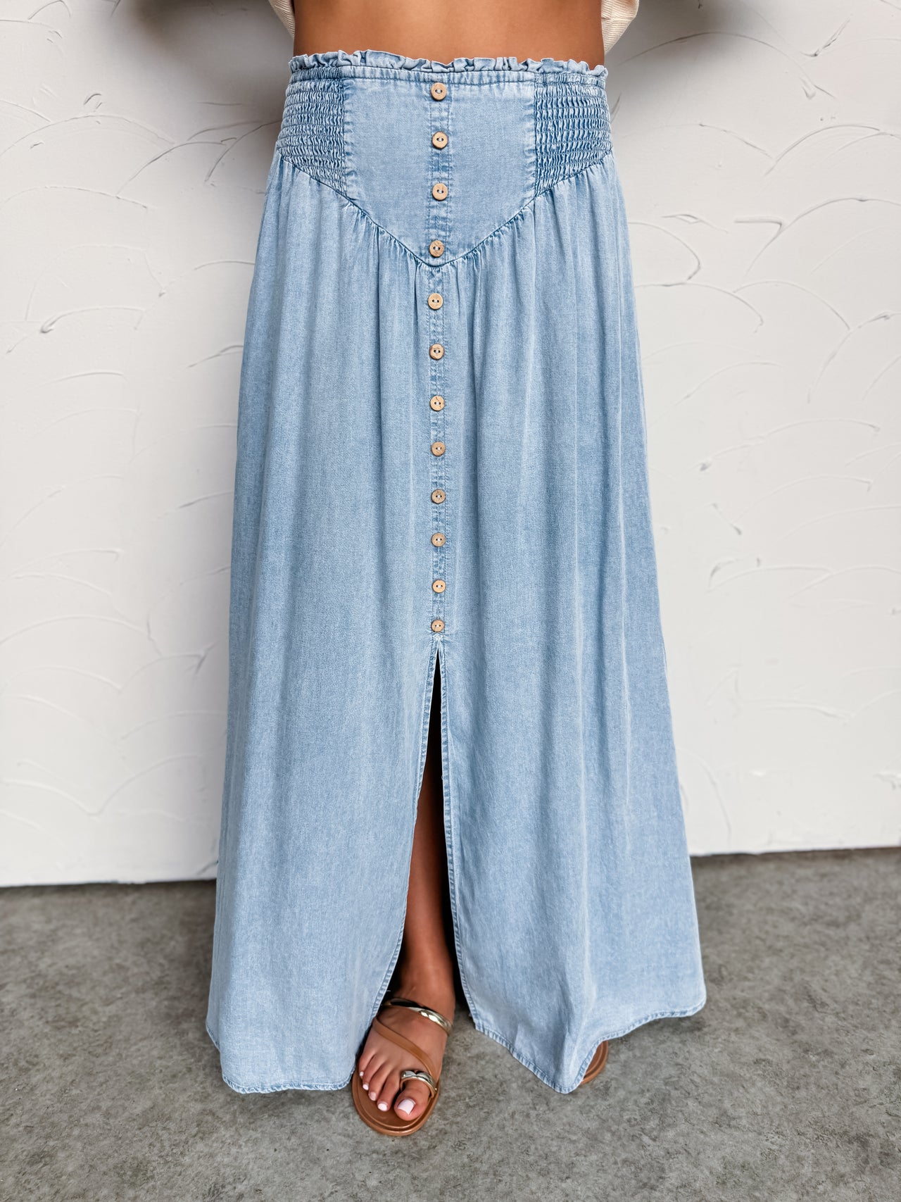 A close-up photograph focusing from the waist down of a woman wearing a high-waisted, light-wash denim maxi skirt made from 100% Tencel fabric against a textured white wall. The skirt features a stretchy smocked waistband, a V-shaped front yoke, a decorative row of faux wooden buttons down the center, and a front slit showing her sandaled foot.