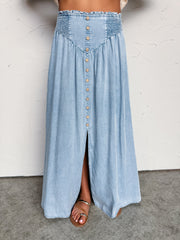 A close-up photograph focusing from the waist down of a woman wearing a high-waisted, light-wash denim maxi skirt made from 100% Tencel fabric against a textured white wall. The skirt features a stretchy smocked waistband, a V-shaped front yoke, a decorative row of faux wooden buttons down the center, and a front slit showing her sandaled foot.