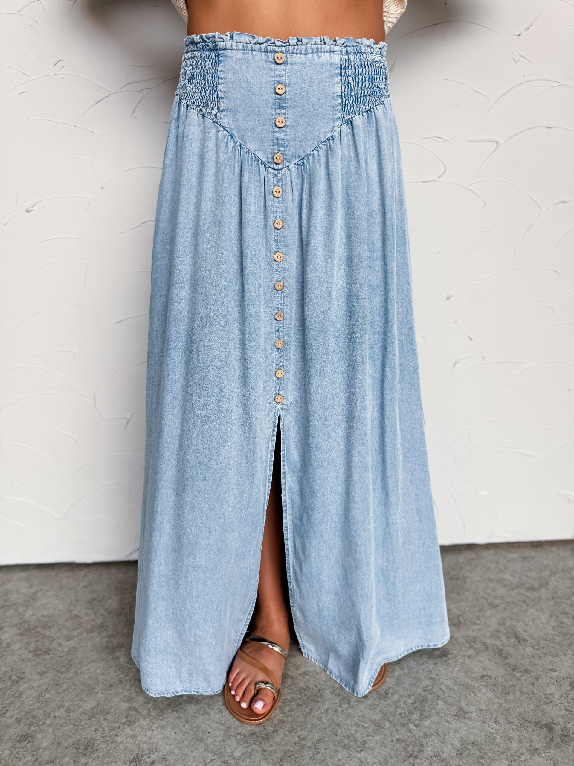A close-up photograph focusing from the waist down of a woman wearing a high-waisted, light-wash denim maxi skirt made from 100% Tencel fabric against a textured white wall. The skirt features a stretchy smocked waistband, a V-shaped front yoke, a decorative row of faux wooden buttons down the center, and a front slit showing her sandaled foot.