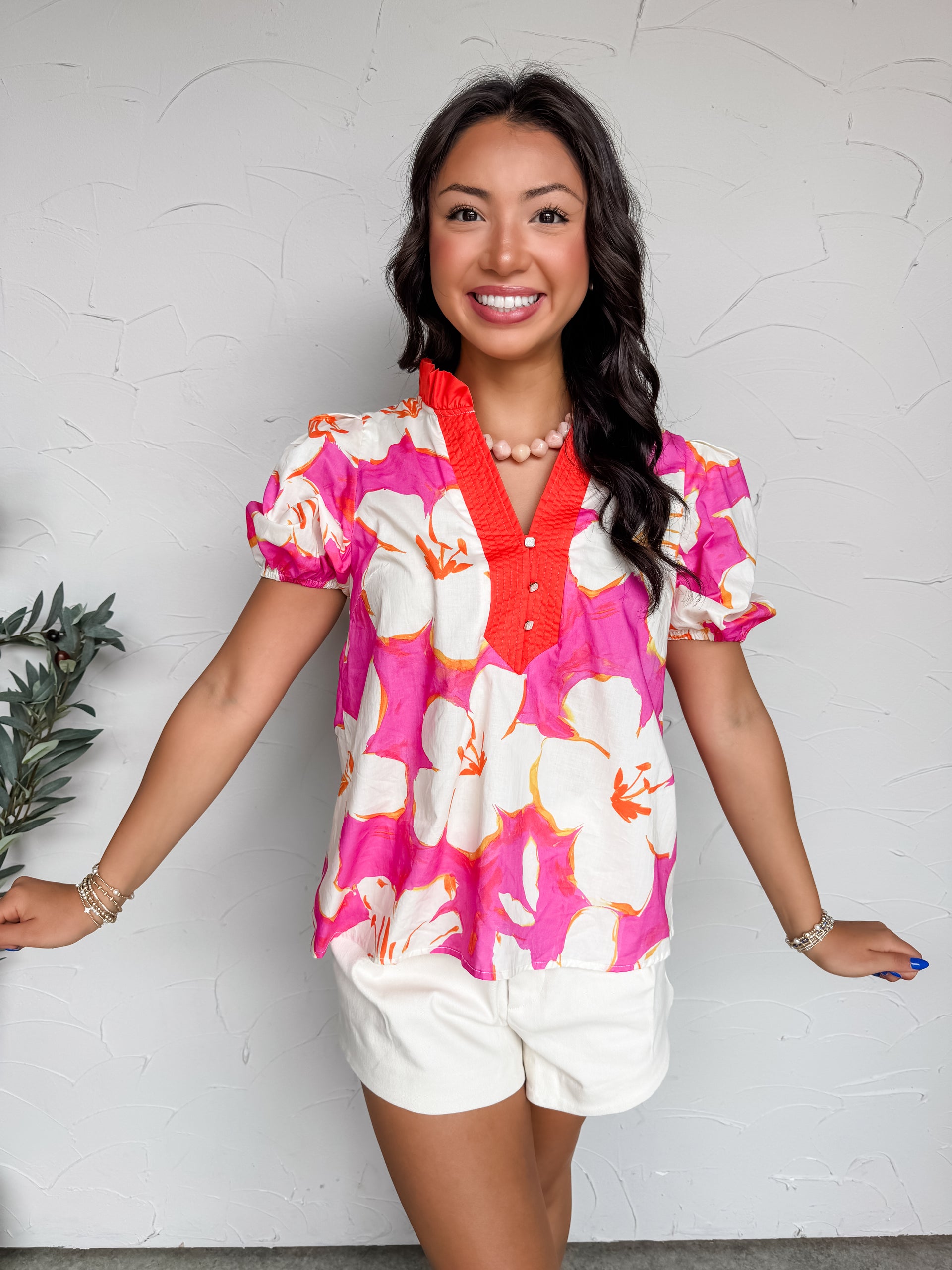 Woman wearing a colorful floral blouse and white shorts against a white wall.
