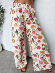 A close-up photograph focusing from the waist down of a woman wearing high-waisted, wide-leg cream pants featuring a colorful pink, orange, and green floral print made from 100% cotton fabric against a textured white wall. The pants are styled with an elastic waistband and show her sandaled feet at the bottom.