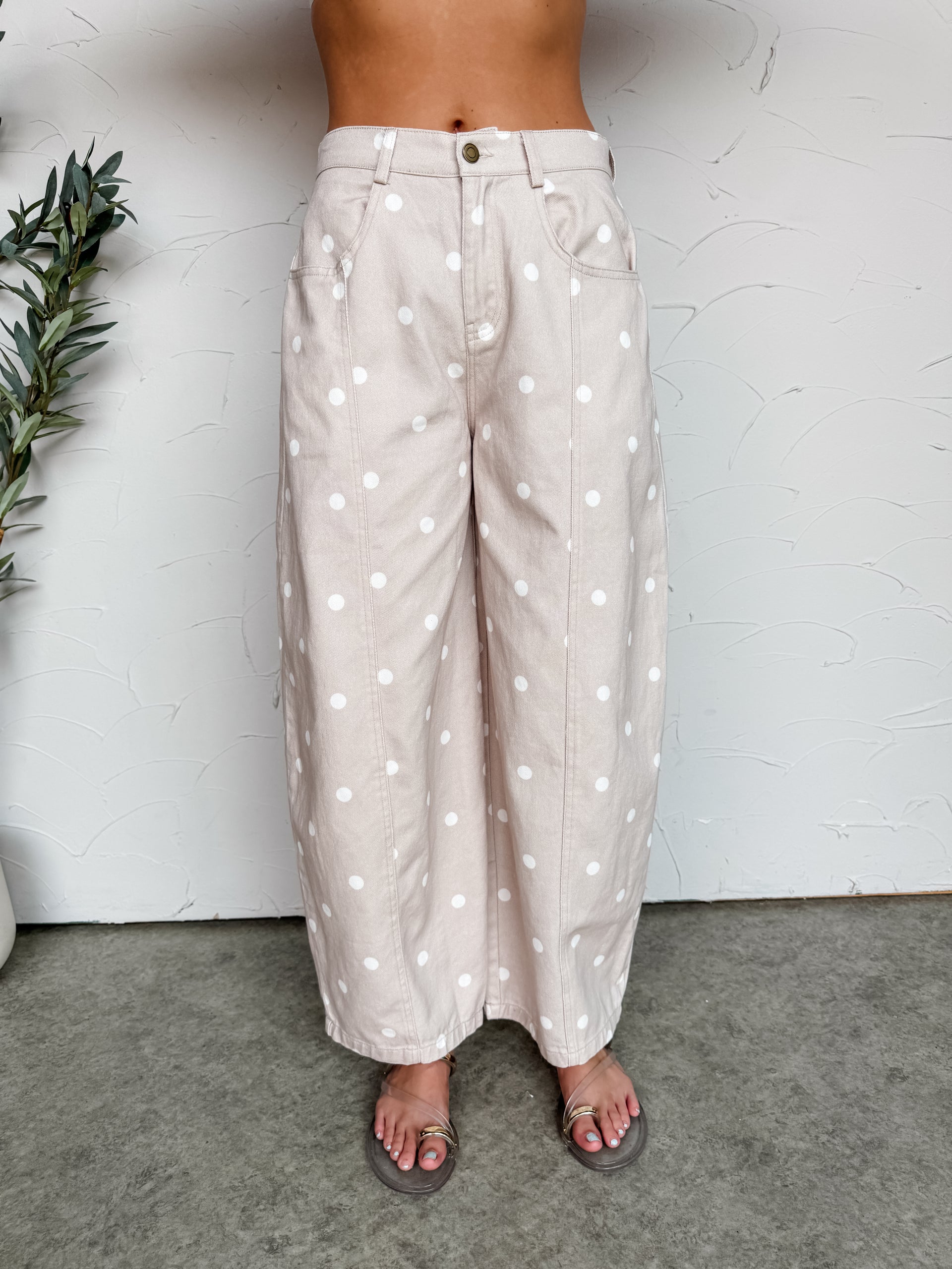 A close-up photograph focusing from the waist down of a woman wearing high-waisted, wide-leg beige pants featuring a white polka dot print made from 100% cotton fabric against a textured white wall. The pants are styled with functional side pockets and vertical seam panels down the center of each leg.