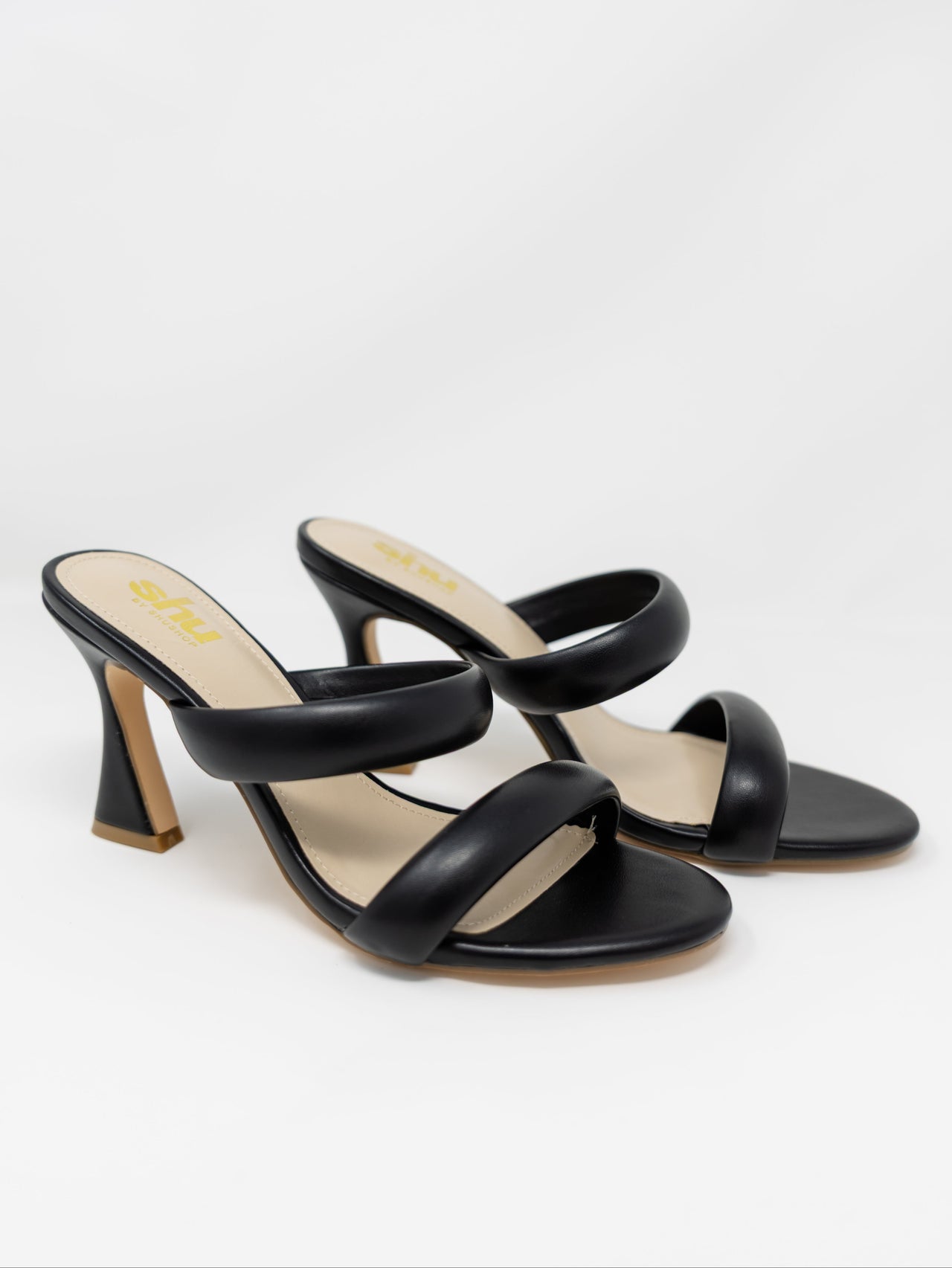 A pair of Shu by Shushop brand black slide-on high heels shown from a side angle against a crisp white background. The open-toe shoes feature two thick, rounded padded black straps across the foot, light beige insoles with a yellow "shu by shushop" logo, and a modern flared high heel.