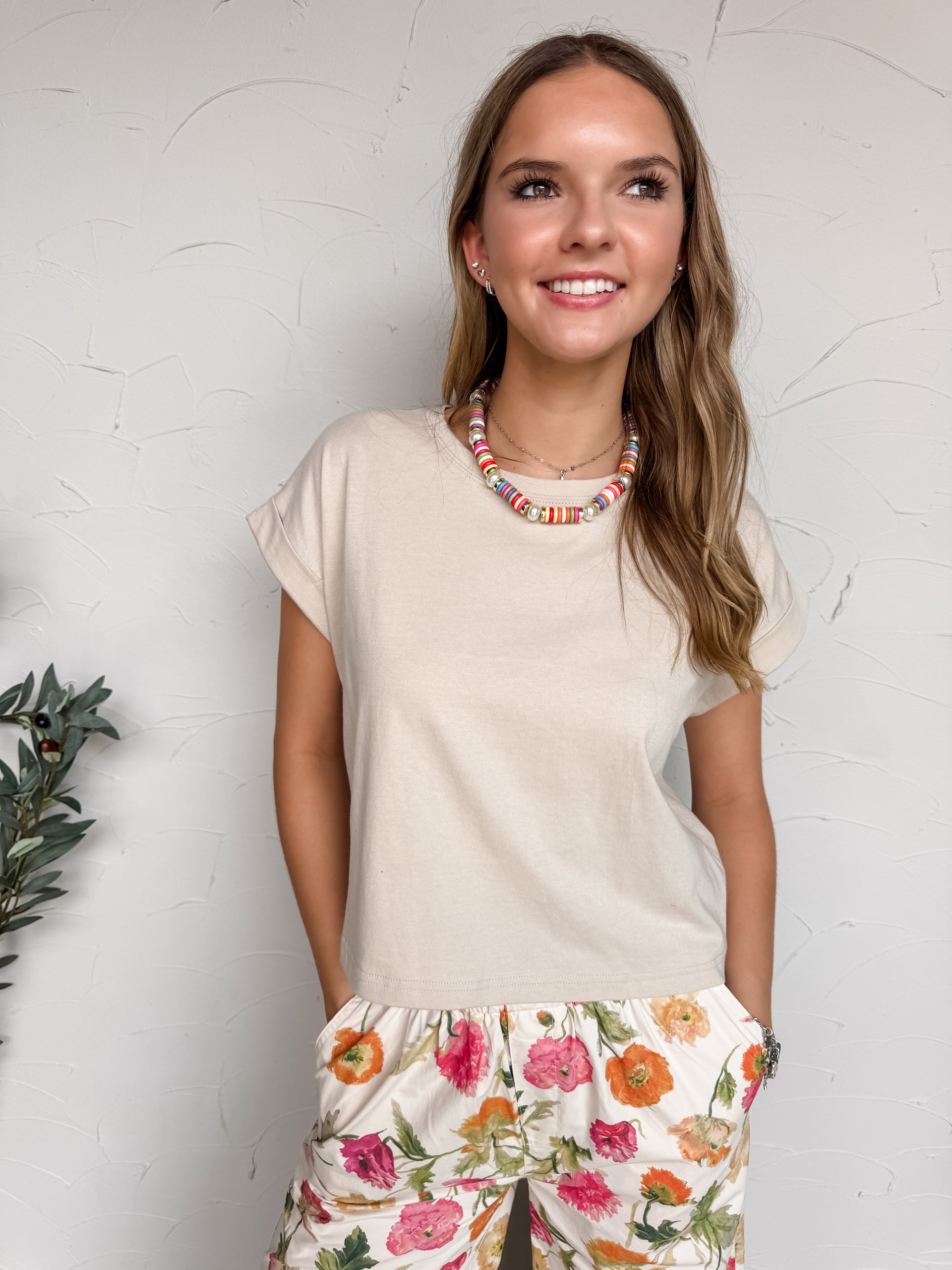 A mid-shot photograph of a smiling blonde woman posing against a textured white wall with her hands in her pockets. She is wearing a neutral beige short-sleeve tee made from 100% cotton fabric, featuring a crew neckline and cuffed cap sleeves, styled with colorful floral-print pants.