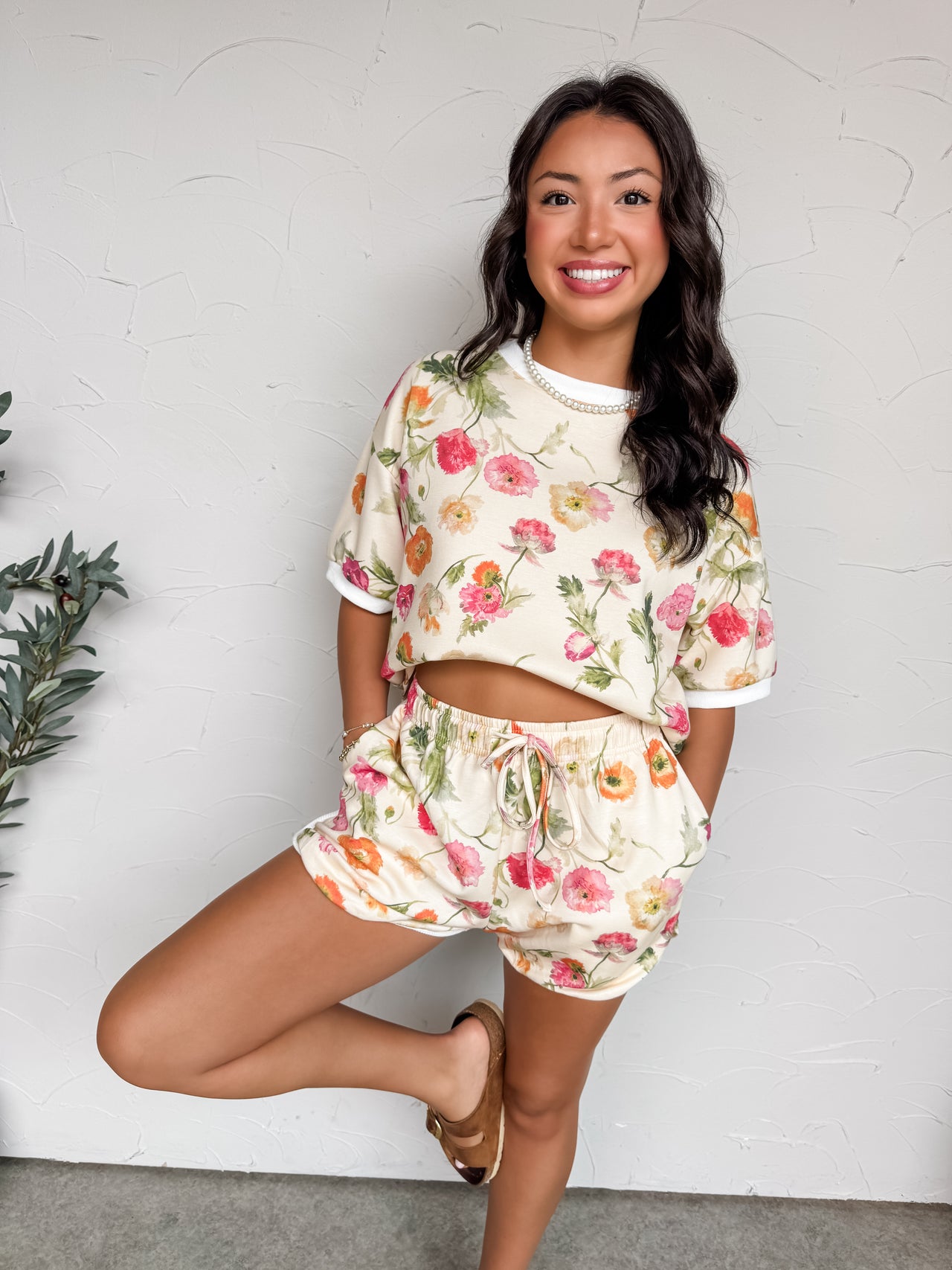 A full-length photograph of a smiling brunette woman posing on one leg against a textured white wall with her hands in her pockets. She is wearing a matching two-piece lounge set made from a soft 50% rayon, 45% polyester, and 5% spandex blend fabric featuring a colorful watercolor floral print, an oversized ringer-style t-shirt, and matching high-waisted drawstring shorts.