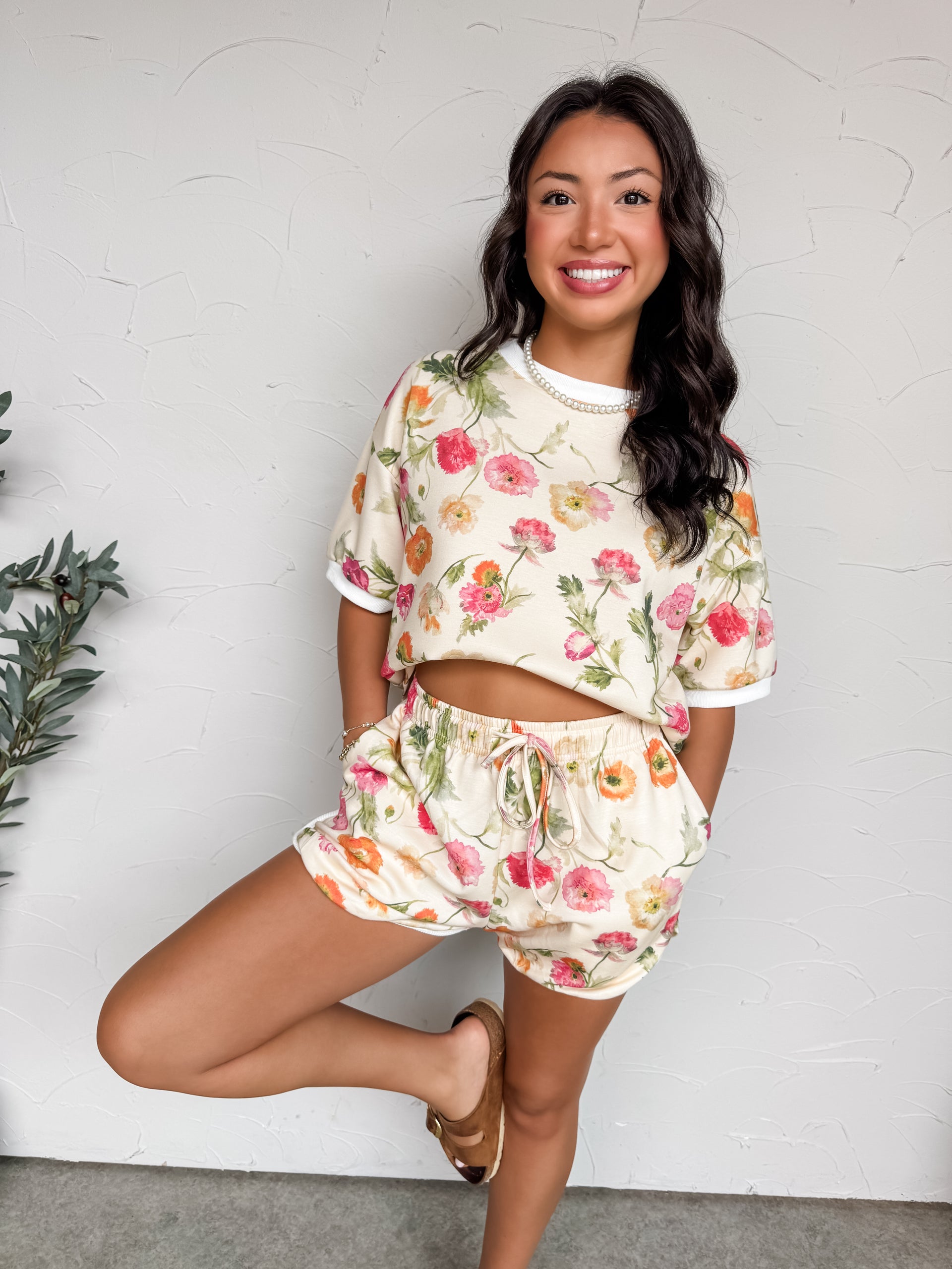 A full-length photograph of a smiling brunette woman posing on one leg against a textured white wall with her hands in her pockets. She is wearing a matching two-piece lounge set made from a soft 50% rayon, 45% polyester, and 5% spandex blend fabric featuring a colorful watercolor floral print, an oversized ringer-style t-shirt, and matching high-waisted drawstring shorts.