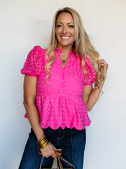 A mid-shot photograph of a smiling blonde woman posing while touching her hair against a textured white wall. She is wearing a vibrant hot pink short-sleeve peplum top made from a 100% cotton eyelet lace fabric, featuring puff sleeves, a ruffled collar, a button-down bodice, and a scalloped peplum hem, styled over dark-wash denim jeans.