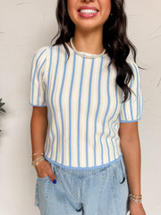 A close-up photograph from the chin down of a smiling brunette woman posing with one hand in her pocket against a textured white wall. She is wearing a short-sleeve knit top made from a soft 50% viscose, 28% polyester, and 22% nylon blend fabric featuring a light blue and cream vertical stripe pattern, subtle puff sleeves, and a crew neckline, styled with light-wash denim shorts.