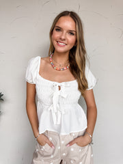 A mid-shot photograph of a smiling blonde woman posing against a textured white wall with her hands in her pockets. She is wearing a sweet white short-sleeve top made from 100% polyester fabric, featuring a smocked bodice, puff sleeves, and a tie-front accent at the neckline, styled with beige and white polka dot pants.