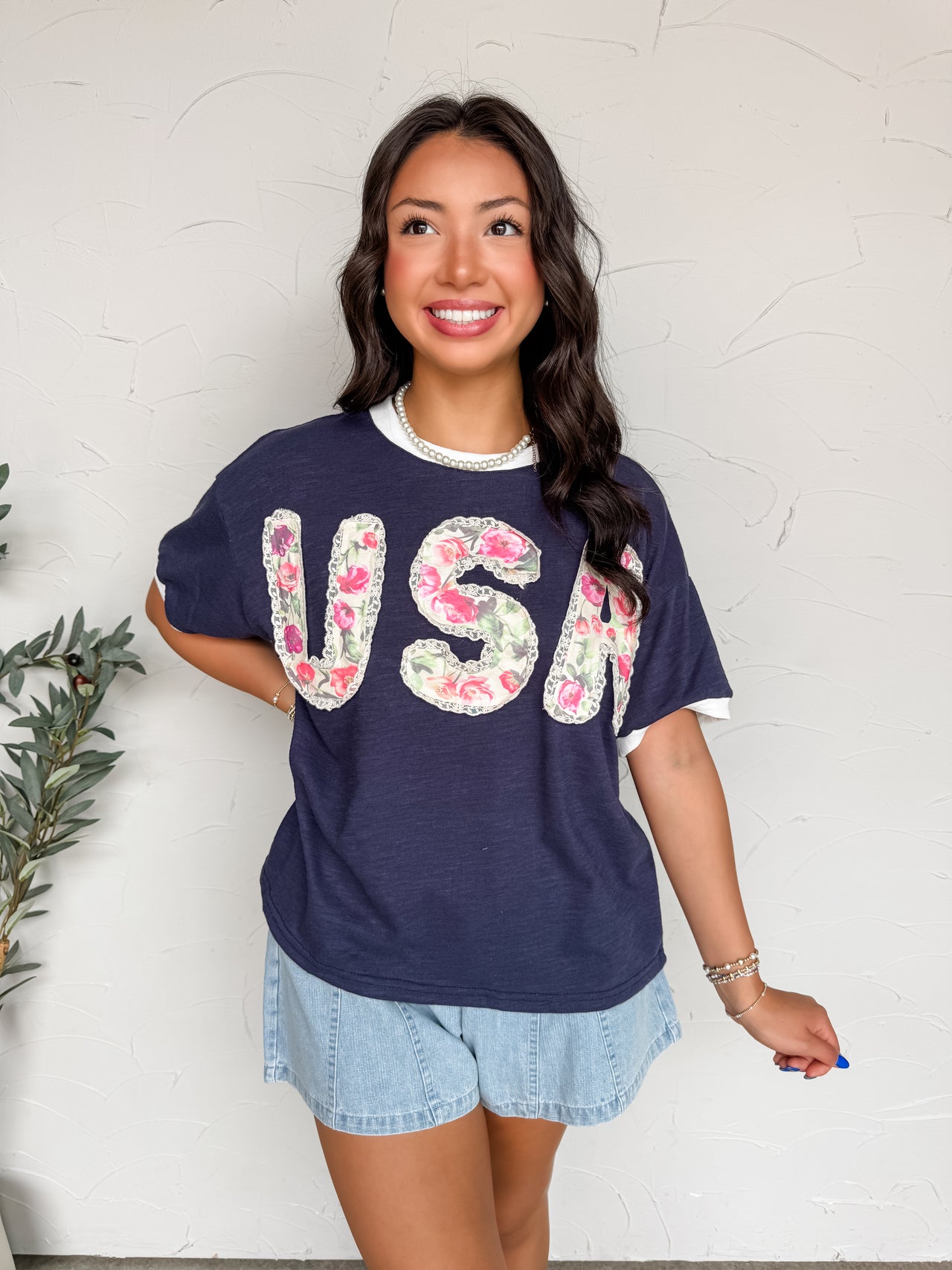 A mid-shot photograph of a smiling brunette woman looking slightly away and posing against a textured white wall. She is wearing a navy blue short-sleeve graphic tee made from a 63% polyester, 35% cotton, and 2% spandex blend fabric featuring "USA" block lettering in a pink floral applique with a lace border, styled with light-wash denim shorts.