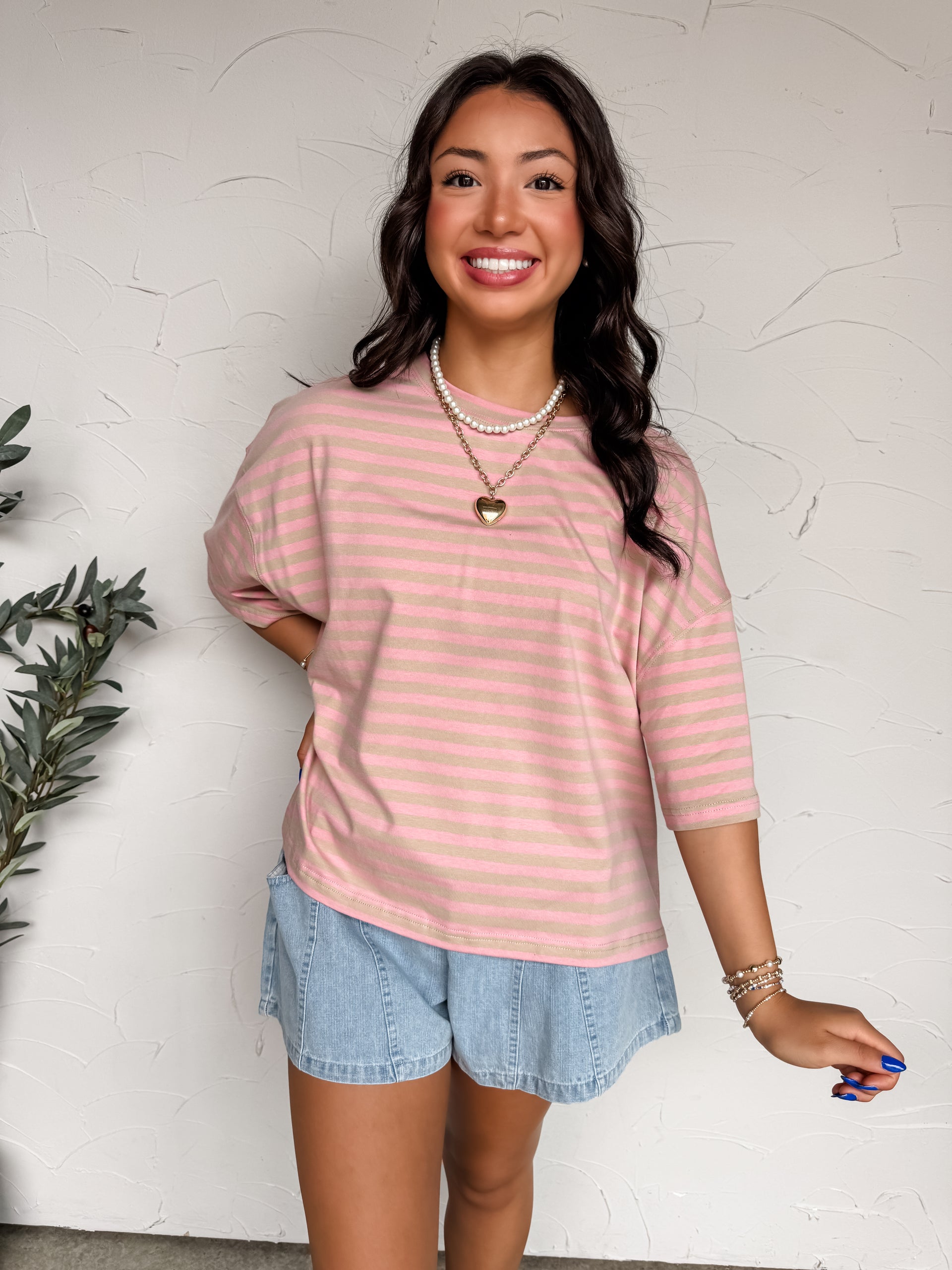 A mid-shot photograph of a smiling brunette woman posing against a textured white wall. She is wearing an oversized crewneck t-shirt made from a 95% cotton and 5% spandex blend fabric featuring a pink and tan horizontal stripe pattern, dropped shoulders, and extended short sleeves, styled with light-wash denim shorts.