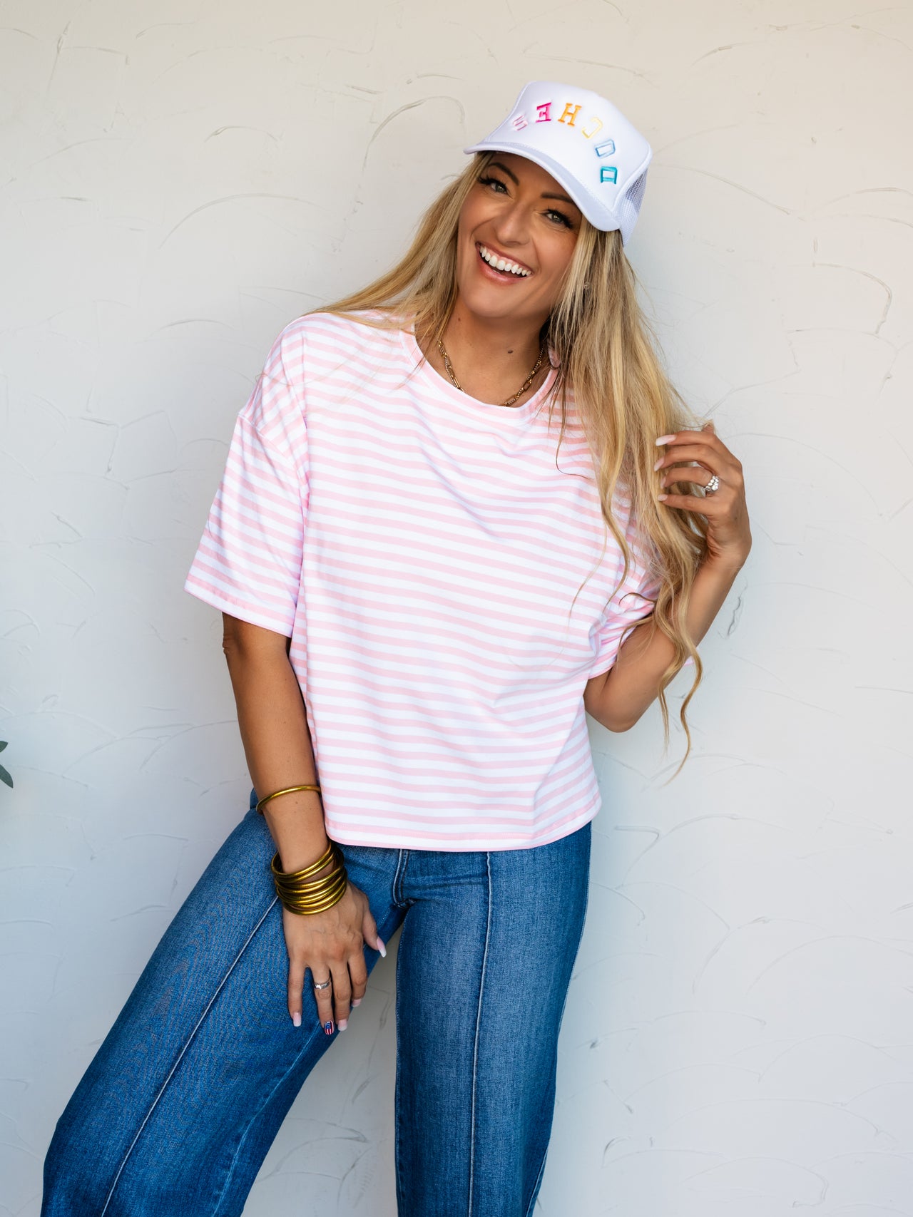 A mid-shot photograph of a laughing blonde woman posing dynamically while touching her hair against a textured white wall. She is wearing a relaxed pink and white horizontally striped short-sleeve t-shirt made from a 95% cotton and 5% spandex blend fabric, styled with dark-wash denim jeans and a white trucker hat with colorful text.