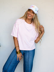 A mid-shot photograph of a laughing blonde woman posing dynamically while touching her hair against a textured white wall. She is wearing a relaxed pink and white horizontally striped short-sleeve t-shirt made from a 95% cotton and 5% spandex blend fabric, styled with dark-wash denim jeans and a white trucker hat with colorful text.