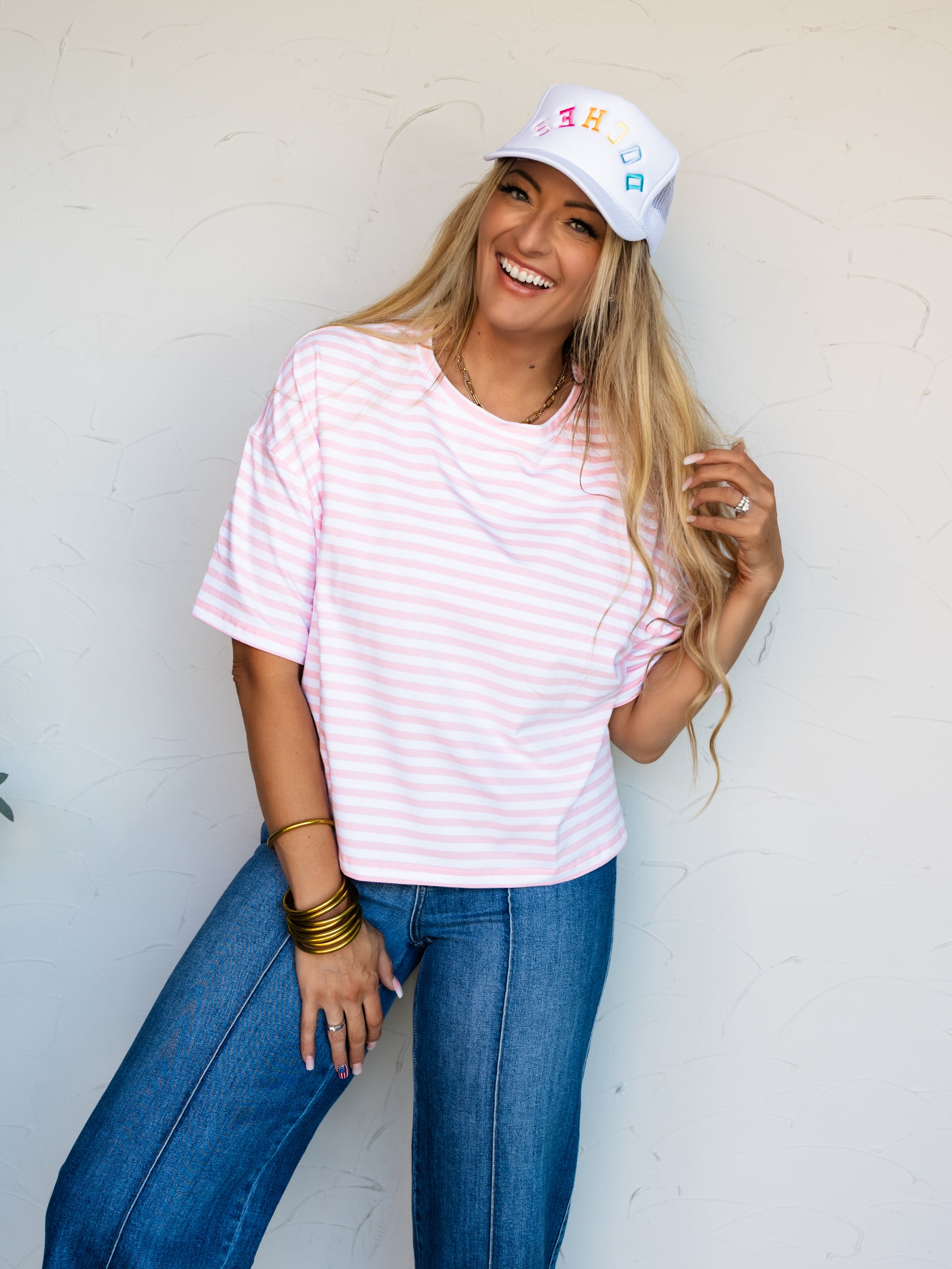 A mid-shot photograph of a laughing blonde woman posing dynamically while touching her hair against a textured white wall. She is wearing a relaxed pink and white horizontally striped short-sleeve t-shirt made from a 95% cotton and 5% spandex blend fabric, styled with dark-wash denim jeans and a white trucker hat with colorful text.