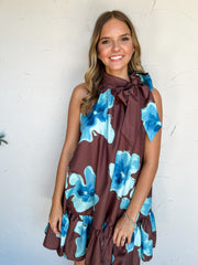 Out Of Town Floral Tie Neck Mini Dress by Glam in 100% polyester, with oversized turquoise and blue florals on deep brown fabric, a side neck bow, and a ruffled hem.
