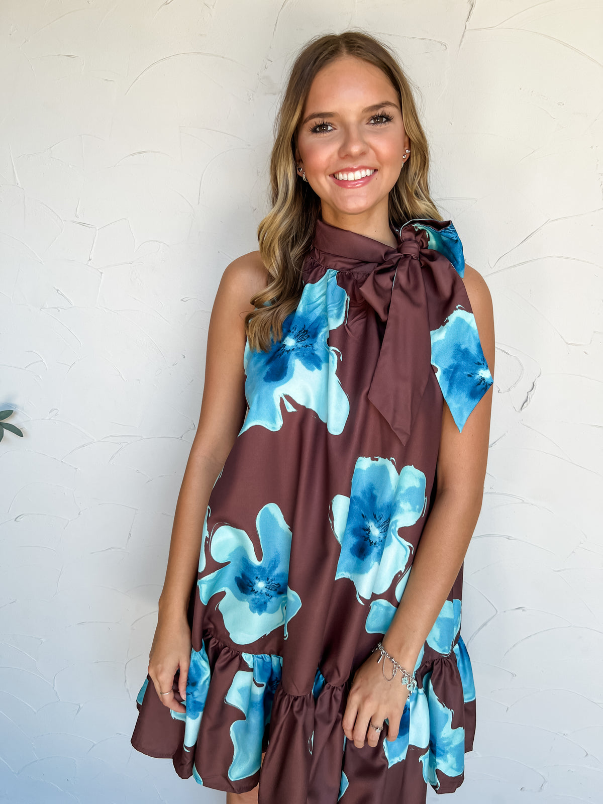 Out Of Town Floral Tie Neck Mini Dress by Glam in 100% polyester, with oversized turquoise and blue florals on deep brown fabric, a side neck bow, and a ruffled hem.
