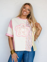 A mid-shot photograph of a smiling blonde woman posing while touching her hair against a textured white wall. She is wearing a relaxed colorblock short-sleeve t-shirt made from an 80% polyester and 20% rayon blend fabric, featuring a cream base panel with a large floral print applique on the chest, bordered by panels of pink, ditsy floral print, and yellow gingham, styled over blue denim.