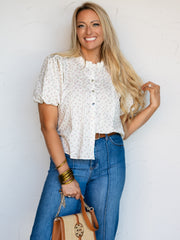 A mid-shot photograph of a smiling blonde woman posing while touching her hair against a textured white wall. She is wearing a cream-colored short-sleeve button-down blouse made from a 100% cotton fabric featuring a ditsy purple and green floral print, puff sleeves, a ruffled collar, and vertical pintuck details, styled tucked into blue denim jeans with a brown belt while holding a woven handbag.