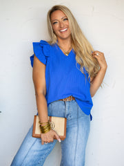 A mid-shot photograph of a smiling blonde woman posing while touching her hair against a textured white wall. She is wearing a royal blue short-sleeve top made from a textured 100% polyester fabric, featuring a V-neckline, a ruffled collar, and layered ruffle sleeves, styled tucked into light-wash blue denim jeans with a brown belt and a woven clutch.