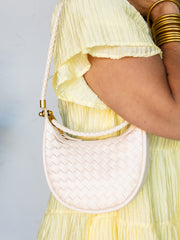 A close-up of a person wearing a pale yellow pleated dress and gold bangles, carrying a small woven cream shoulder bag with a braided strap and gold-tone hardware.