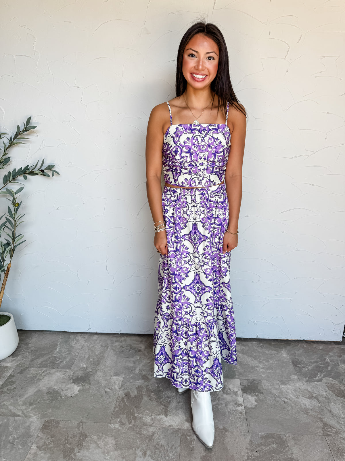 A full-length photograph of a smiling brunette woman standing against a textured white wall next to a potted olive plant in a white pot. She is wearing a two-piece matching set made from 100% cotton in a purple and white Mediterranean tile print, consisting of a sleeveless square-neck crop top with thin cami straps and a high-waisted tiered maxi skirt, paired with pointed-toe white ankle boots, styled with a gold pendant necklace, a gold bracelet, and beaded wrist accessories.