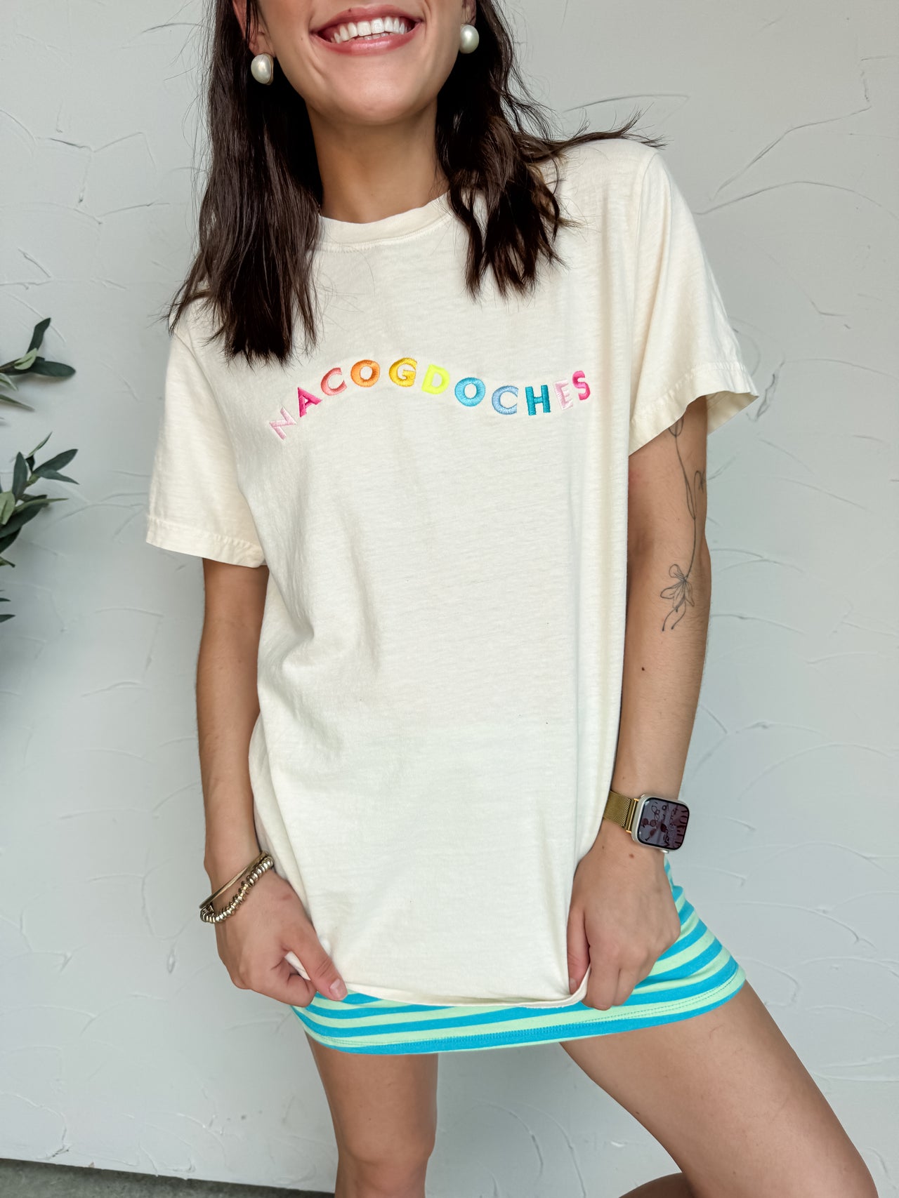A close-up photograph from the nose down of a smiling brunette woman posing against a textured white wall. She is wearing an oversized cream short-sleeve t-shirt featuring the town name "Nacogdoches" embroidered across the front in a colorful arched rainbow font, styled over a bright blue and green horizontally striped mini skirt.