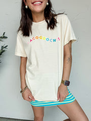 A close-up photograph from the nose down of a smiling brunette woman posing against a textured white wall. She is wearing an oversized cream short-sleeve t-shirt featuring the town name "Nacogdoches" embroidered across the front in a colorful arched rainbow font, styled over a bright blue and green horizontally striped mini skirt.