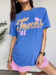 A close-up photograph from the nose down of a brunette woman posing against a textured white wall. She is wearing an oversized periwinkle blue short-sleeve t-shirt featuring retro-style "Texas '45" graphics printed across the front in tan, orange, and pink, styled over pink and white gingham shorts.