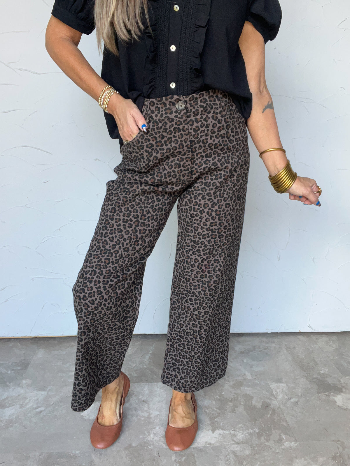 Model wearing Speak The Truth Wide Leg Leopard Pants by Umgee, made from 97% cotton and 3% spandex, featuring an allover dark brown and black leopard print on a taupe background, a front button closure, front pockets, and cropped wide legs, styled with a black ruffled button-front blouse and brown shoes.


