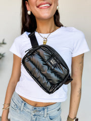 A close-up shot of a person smiling and wearing a Lulu Mac brand black puffer belt bag across their chest as a crossbody. The bag features a glossy, quilted black puffer fabric design, a top zipper closure, an adjustable black webbed strap, and a small black patch in the center with the words "LULU MAC" in white text. The person is wearing a white t-shirt, light-wash denim jeans, and a gold heart necklace.
