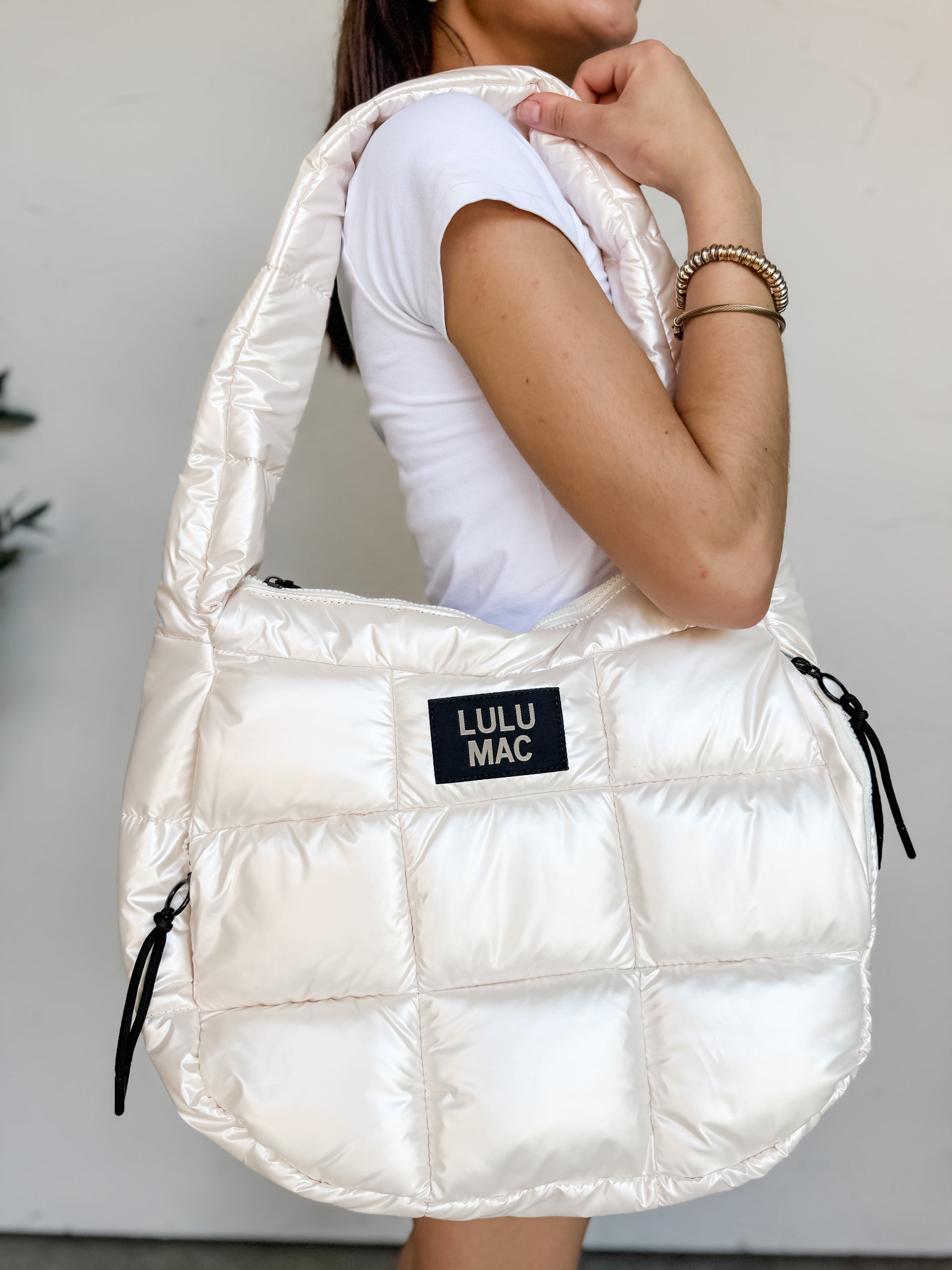A person holding a large Lulu Mac brand puffer tote bag over their shoulder. The bag is made from a glossy, pearlescent off-white quilted fabric with a cloud-like texture. It features a wide, matching puffer shoulder strap, black zipper pull cords on the sides, and a black rectangular patch in the center with the words "LULU MAC" in white text. The person is wearing a white t-shirt and gold bracelets.