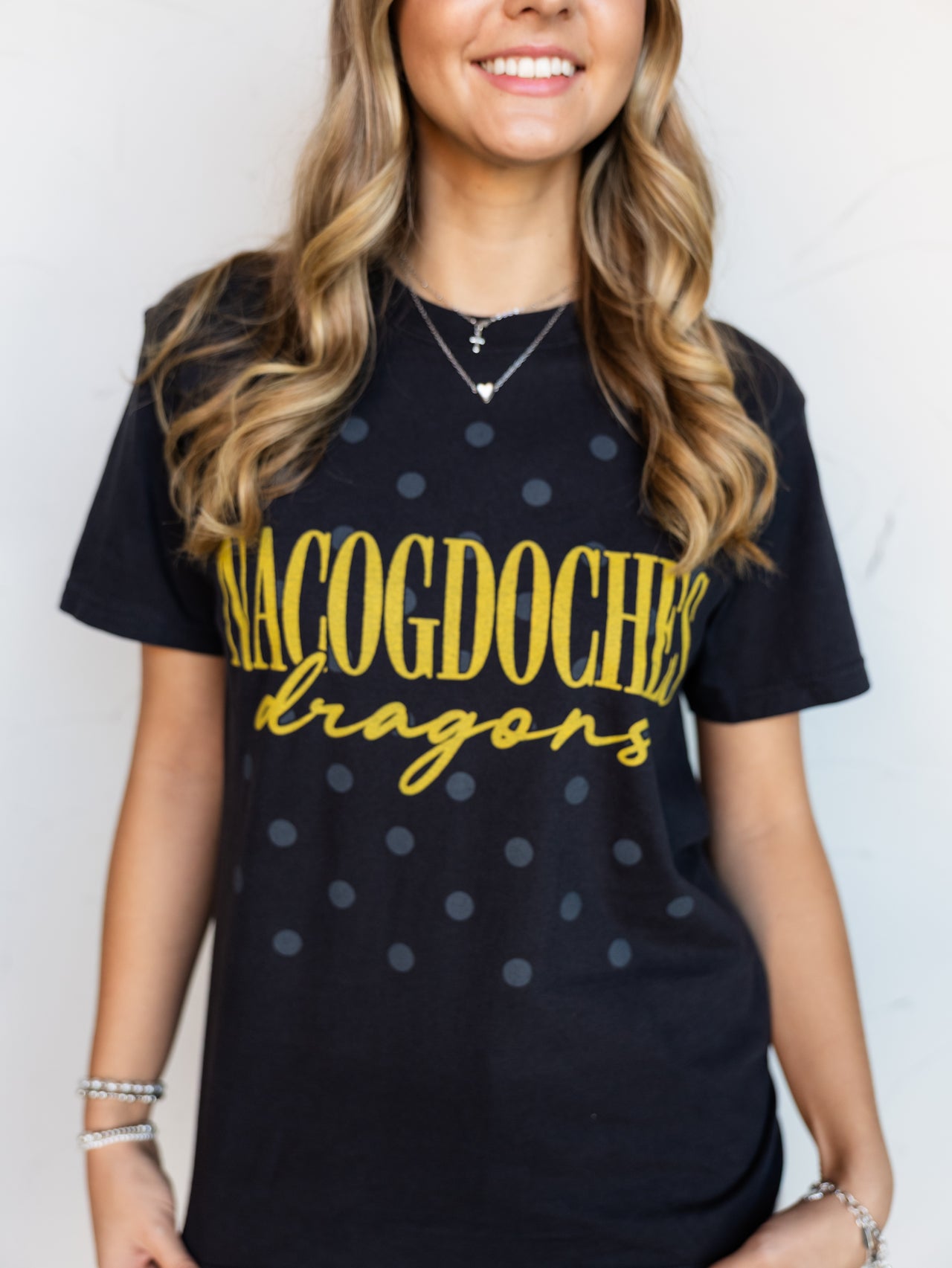 A close-up shot of a smiling person wearing a black crewneck t-shirt with a subtle, all-over dark gray polka dot pattern. The shirt features the word "NACOGDOCHES" printed in tall gold block letters with "dragons" written underneath in a matching gold cursive script font. The person is accessorized with silver layered necklaces and bracelets.