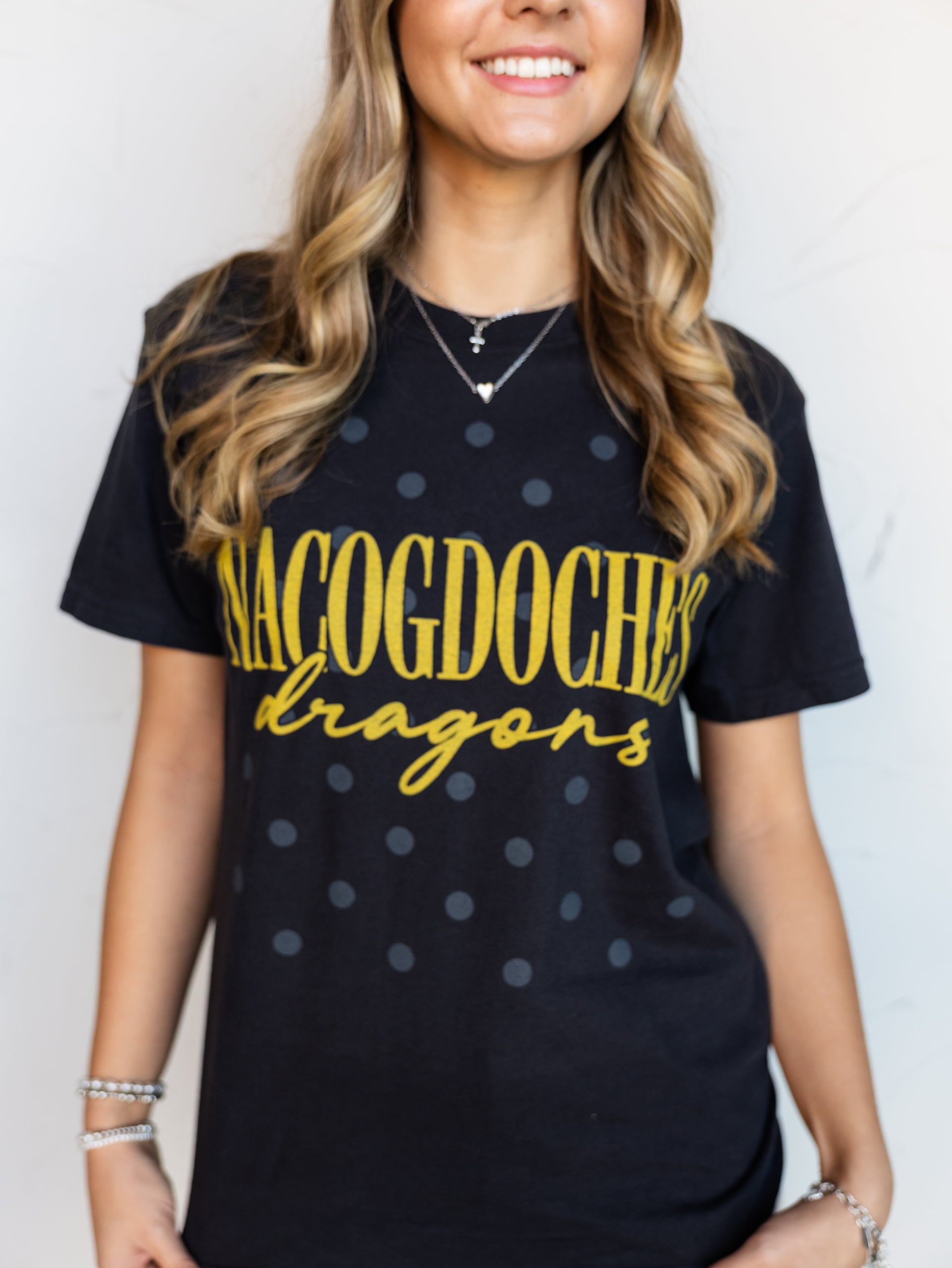 A close-up shot of a smiling person wearing a black crewneck t-shirt with a subtle, all-over dark gray polka dot pattern. The shirt features the word "NACOGDOCHES" printed in tall gold block letters with "dragons" written underneath in a matching gold cursive script font. The person is accessorized with silver layered necklaces and bracelets.