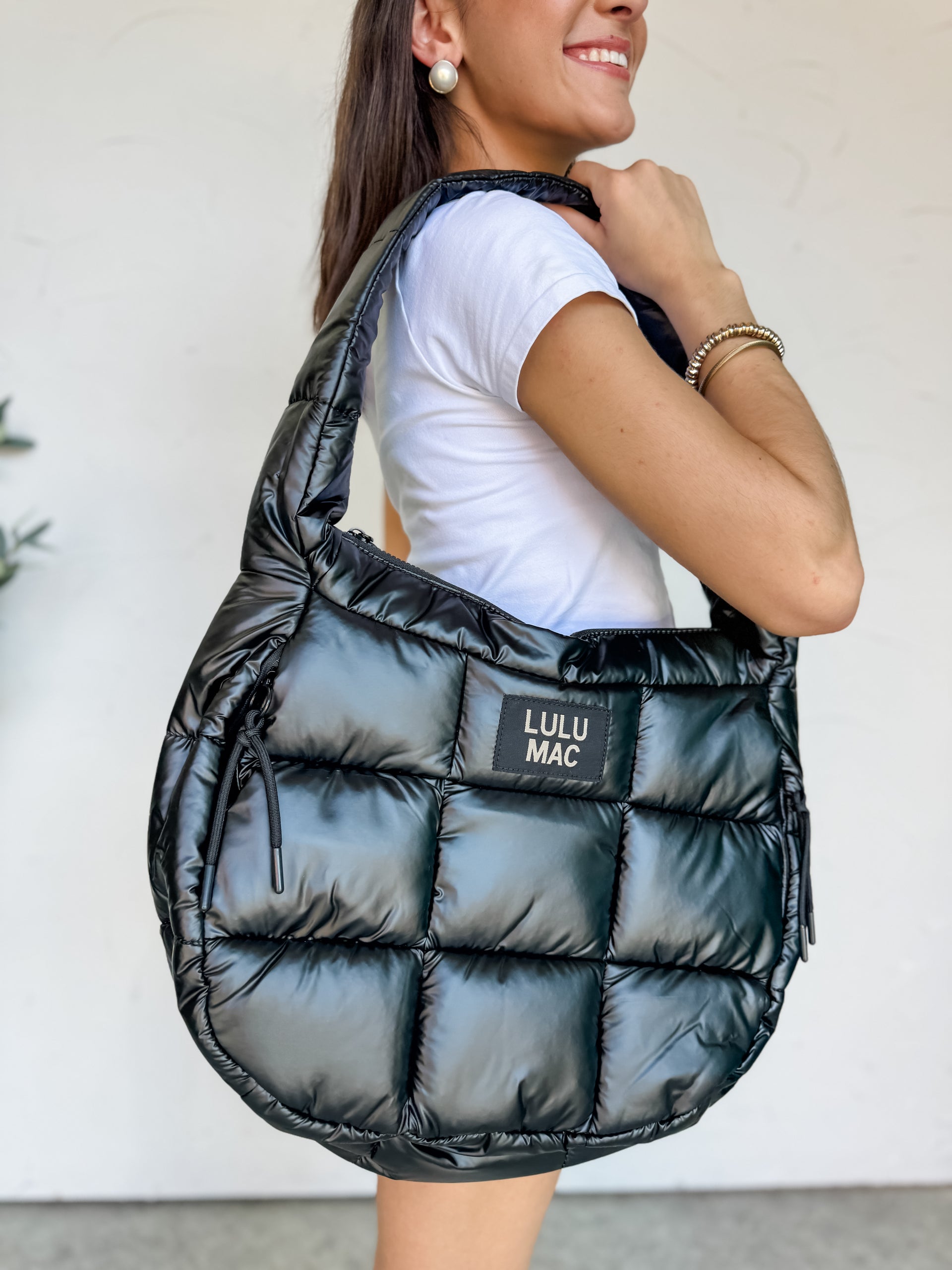 A close-up shot of a person smiling and holding a large Lulu Mac brand puffer tote bag over their shoulder. The bag is made from a high-shine, glossy black quilted fabric with a padded texture. It features a wide, matching black puffer shoulder strap, sleek zipper pull cords on the sides, and a black rectangular patch in the center with the words "LULU MAC" in white text. The person is wearing a white t-shirt and gold bracelets.