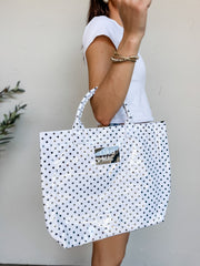 A person carrying a Lulu Mac brand large tote bag with a glossy finish. The bag features a white background covered in a pattern of small black polka dots, matching structured top handles, and a reflective silver metallic rectangular patch in the center with the words "LULU MAC" printed on it. The person holding the bag is wearing a white t-shirt and gold bracelets.