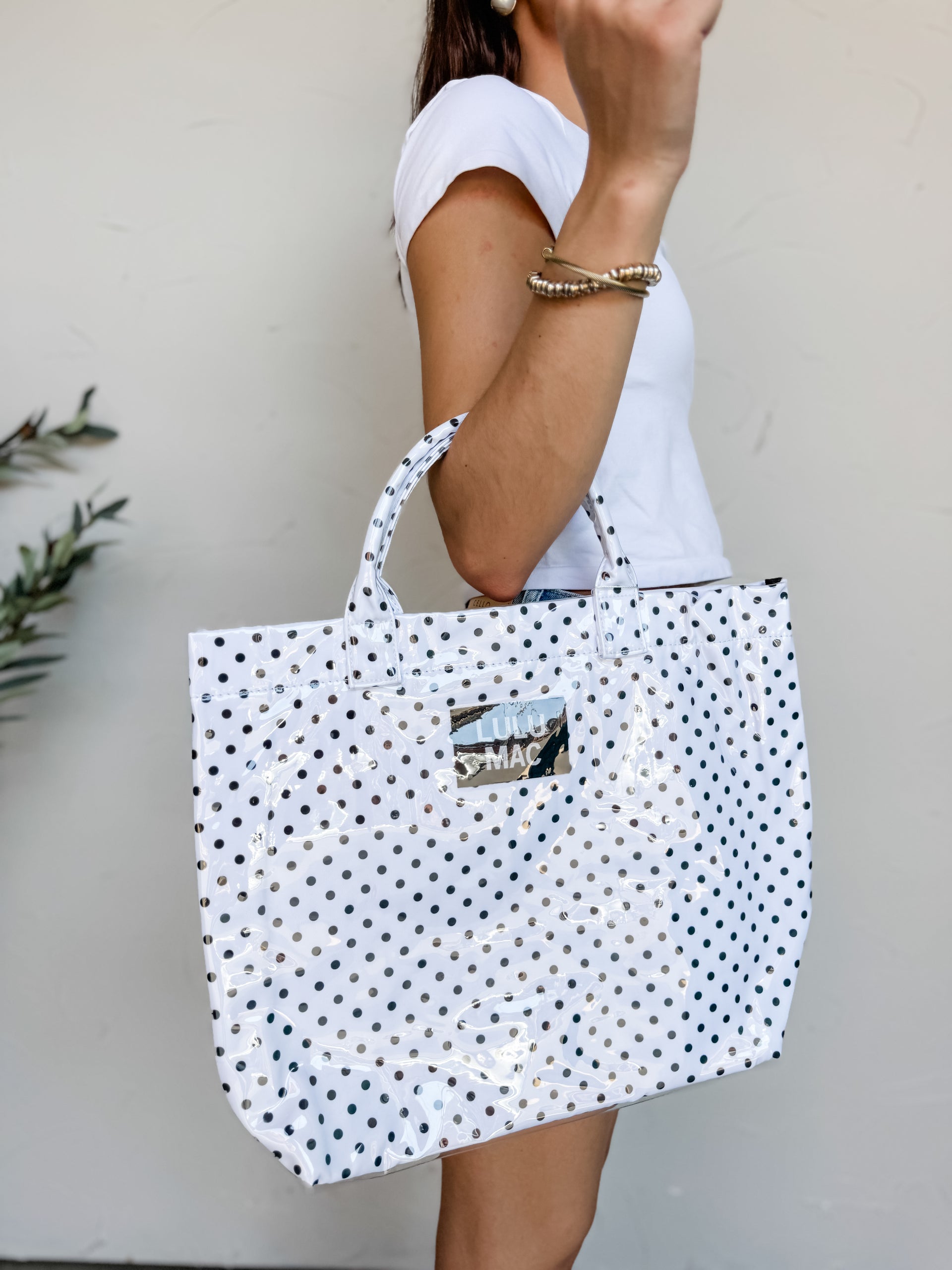 A person carrying a Lulu Mac brand large tote bag with a glossy finish. The bag features a white background covered in a pattern of small black polka dots, matching structured top handles, and a reflective silver metallic rectangular patch in the center with the words "LULU MAC" printed on it. The person holding the bag is wearing a white t-shirt and gold bracelets.