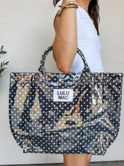 A person carrying a large Lulu Mac brand tote bag with a high-gloss finish. The bag features a black background covered in a pattern of small white polka dots, matching black structured top handles, and a rectangular white fabric patch in the center with the words "LULU MAC" printed in black text. The person holding the bag is wearing a white t-shirt and gold beaded bracelets