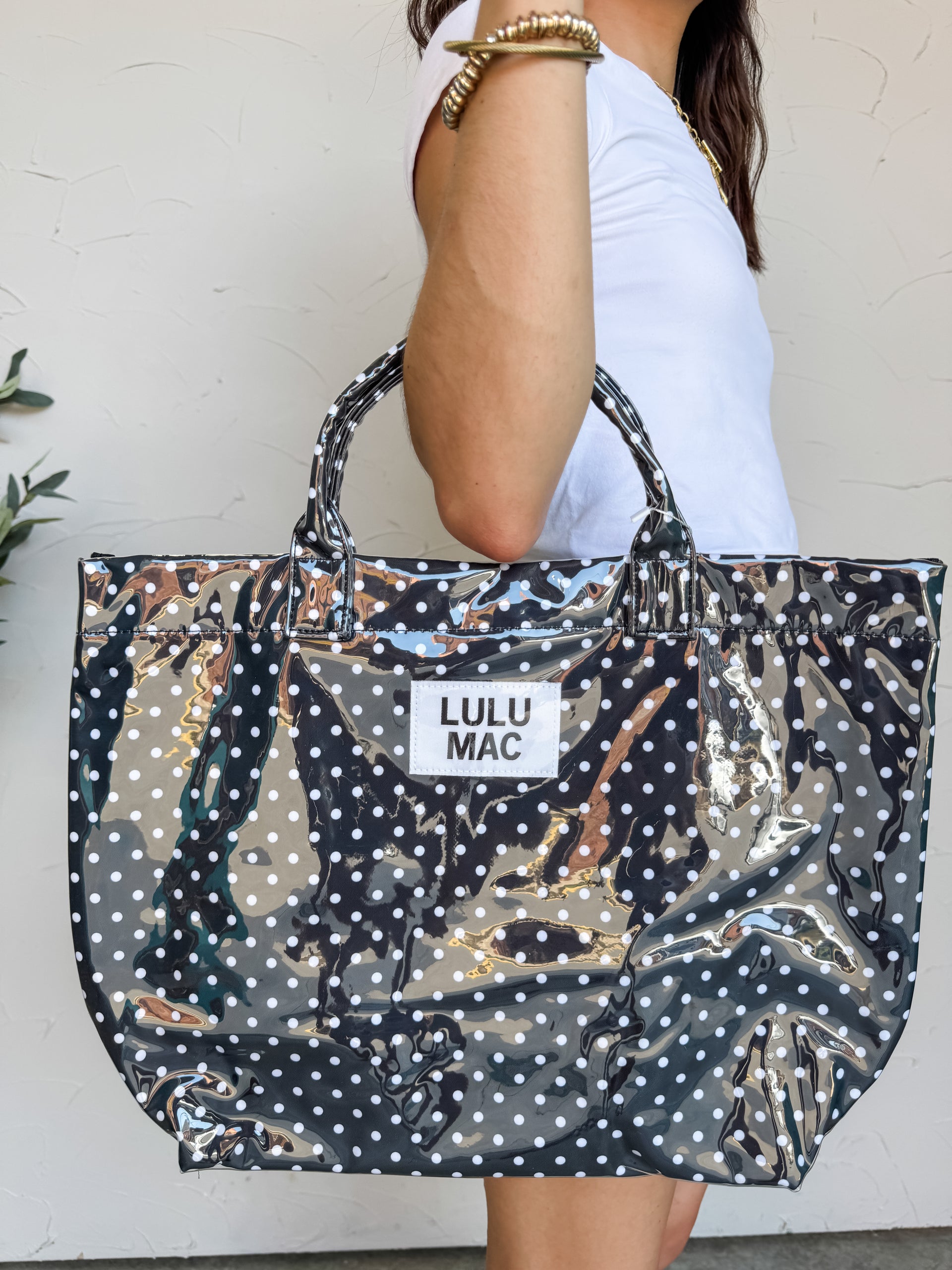 A person carrying a large Lulu Mac brand tote bag with a high-gloss finish. The bag features a black background covered in a pattern of small white polka dots, matching black structured top handles, and a rectangular white fabric patch in the center with the words "LULU MAC" printed in black text. The person holding the bag is wearing a white t-shirt and gold beaded bracelets